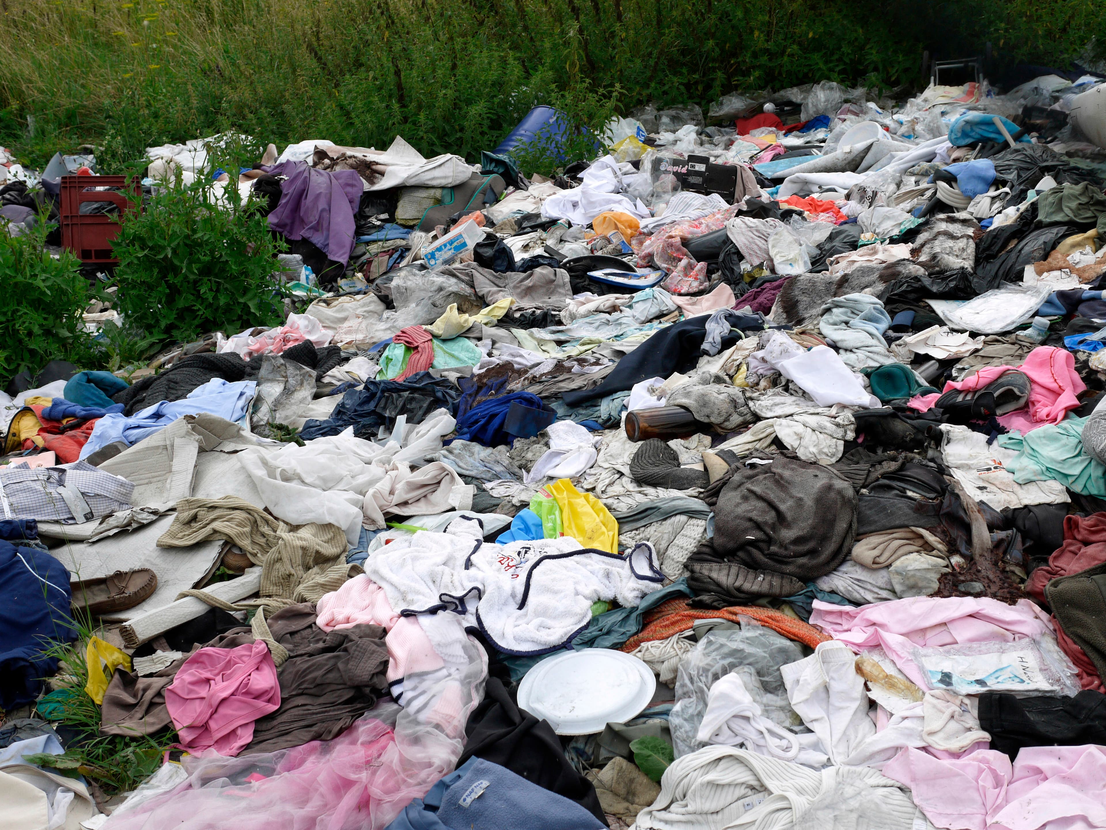 Amoncellement de vêtements usagés abandonnés à même le sol en bordure de végétation, mêlés à des sacs plastiques et à des déchets divers.