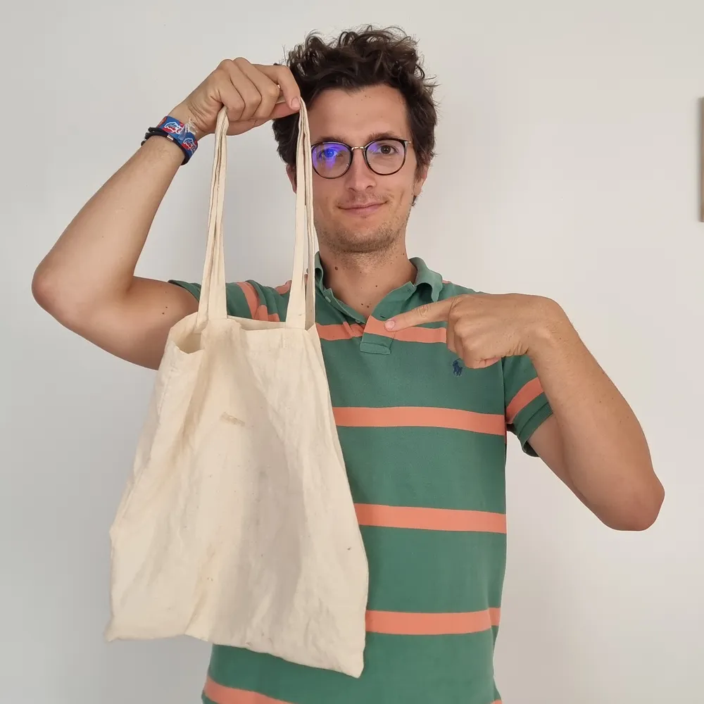 Stanislas tient un tote bag en coton écru à bras levé et le montre du doigt