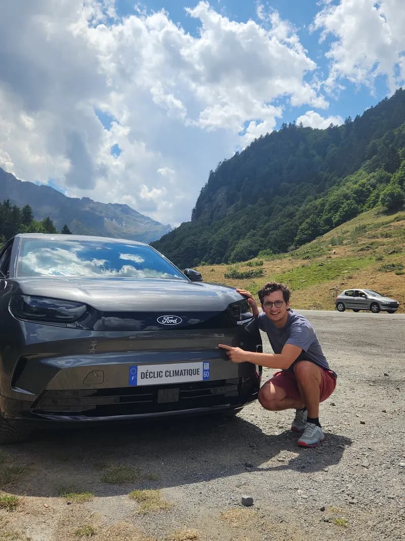 Stanislas Goubault accroupi devant une voiture électrique de location, la main sur sa plaque d'immatriculation personnalisée Déclic Climatique, sur une route de montagne en Espagne